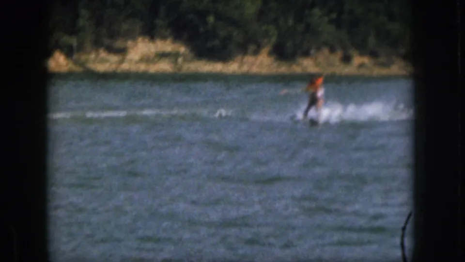 Distant 1961 Michigan water-skiing view with the skier near the right side, white spray behind, and wooded shoreline in the background.