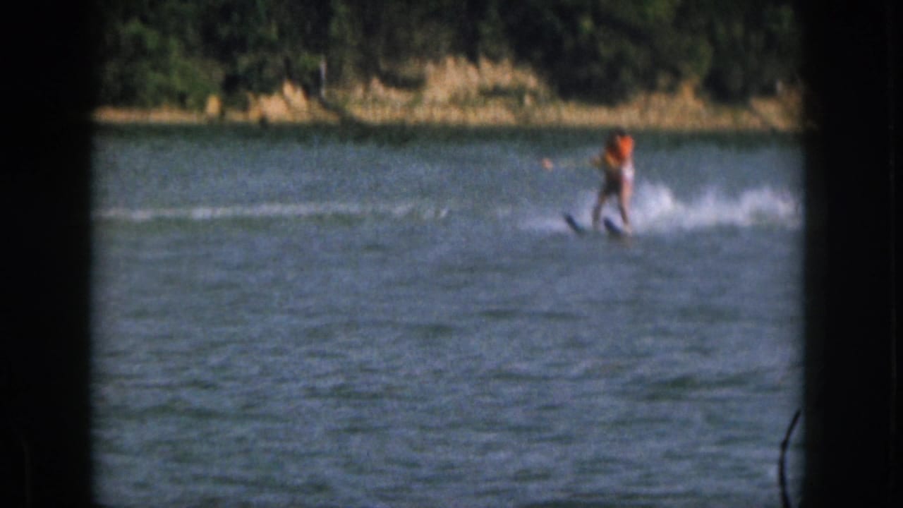 1961 Michigan home-movie frame showing a woman in an orange life vest water skiing across a lake with trees in the distance.