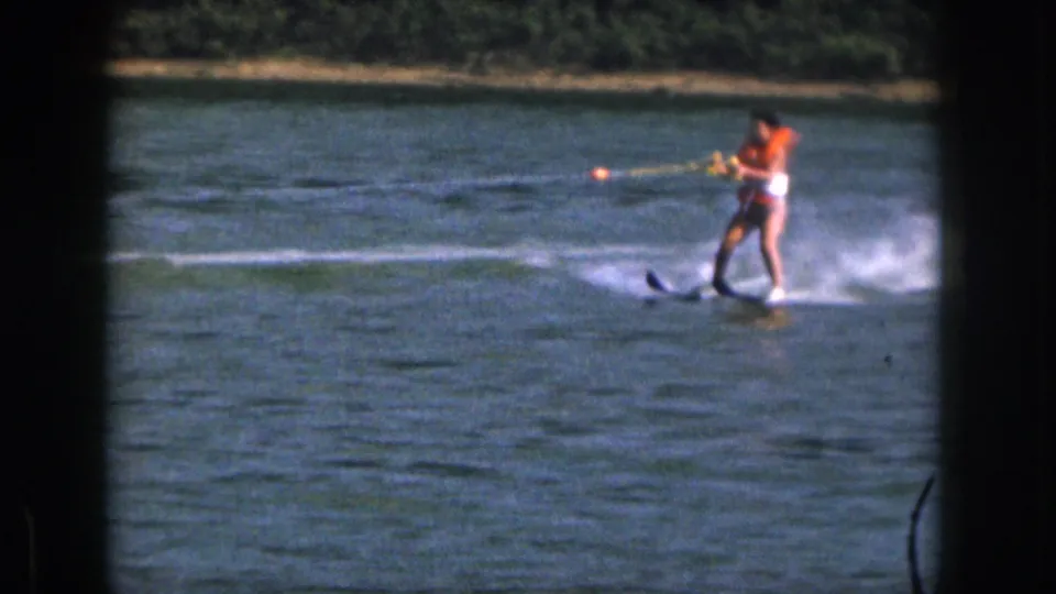 1961 Michigan home-movie still with the woman water skiing nearer the camera, orange vest visible against the lake surface.
