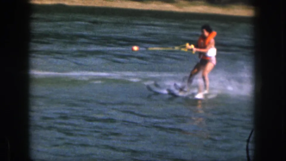 Close 1961 Michigan frame of a woman in an orange life vest water skiing, with tow rope, skis and lake spray clearly visible.