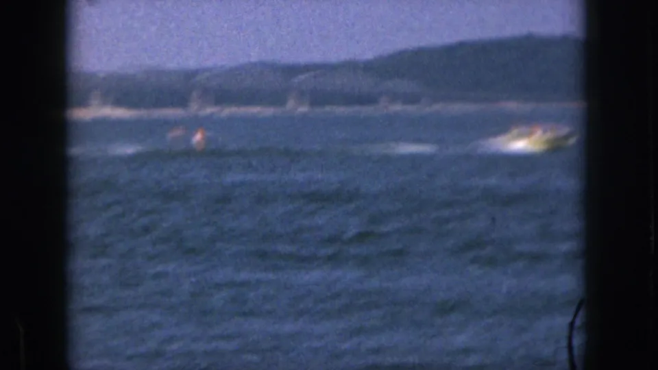 Distant 1961 Michigan lake view with a motorboat towing a water skier across open water near a wooded shoreline.