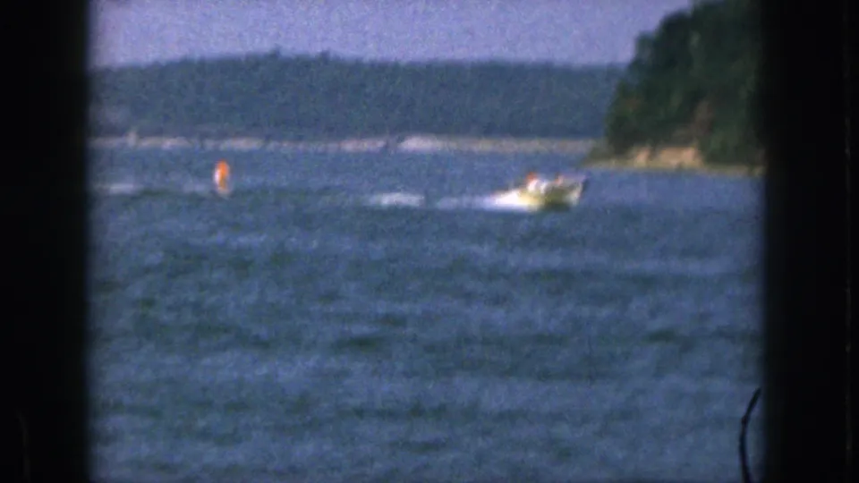 1961 Michigan home-movie frame with a small motorboat towing a skier across blue water near a wooded point.