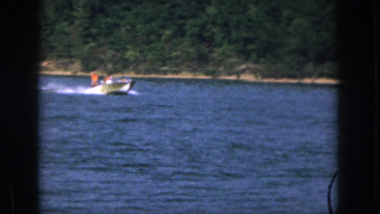Motorboat with a person in orange moving across the lake near a wooded bank.