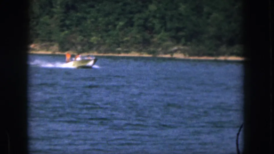 1961 Michigan lake scene with a small motorboat and distant passenger in orange crossing in front of trees.