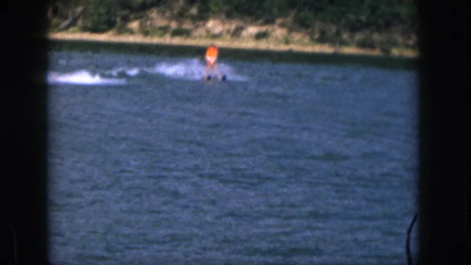 1961 Michigan home-movie frame showing a distant water skier in orange on blue lake water near a wooded shore.