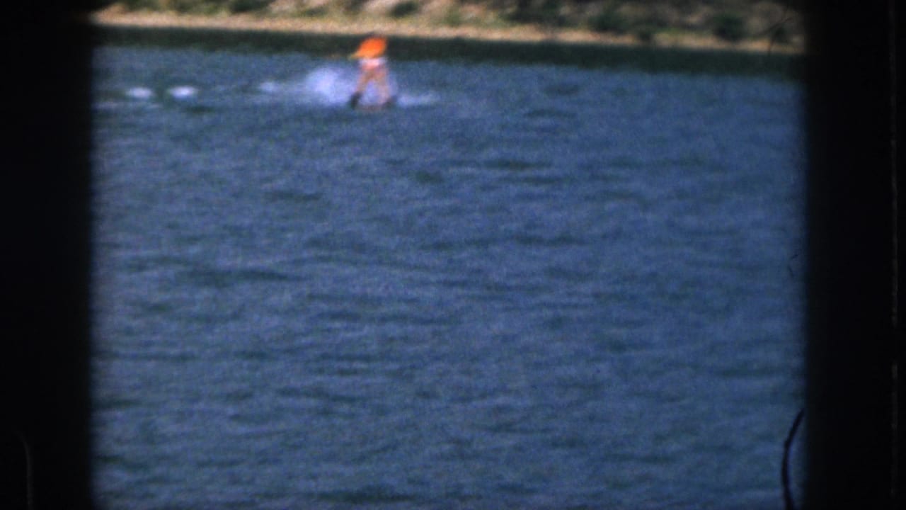 1961 Michigan home-movie frame showing a distant water-skier in an orange life vest moving across a pond with shoreline behind.