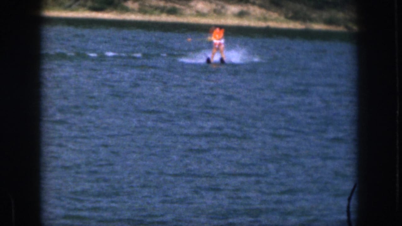 Distant 1961 Michigan water-skiing frame with an orange-vested skier, blue water, white spray, and wooded shoreline.