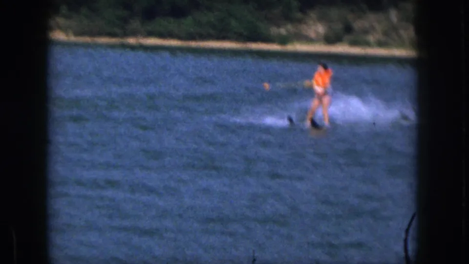 1961 Michigan home-movie still with a water-skier in orange life vest and white trunks kicking up spray on the pond.
