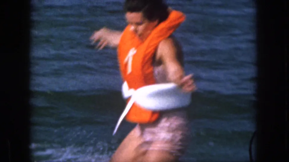 The home-movie frame shows the woman close to the camera as she falls or regains balance in the water while wearing an orange life vest.