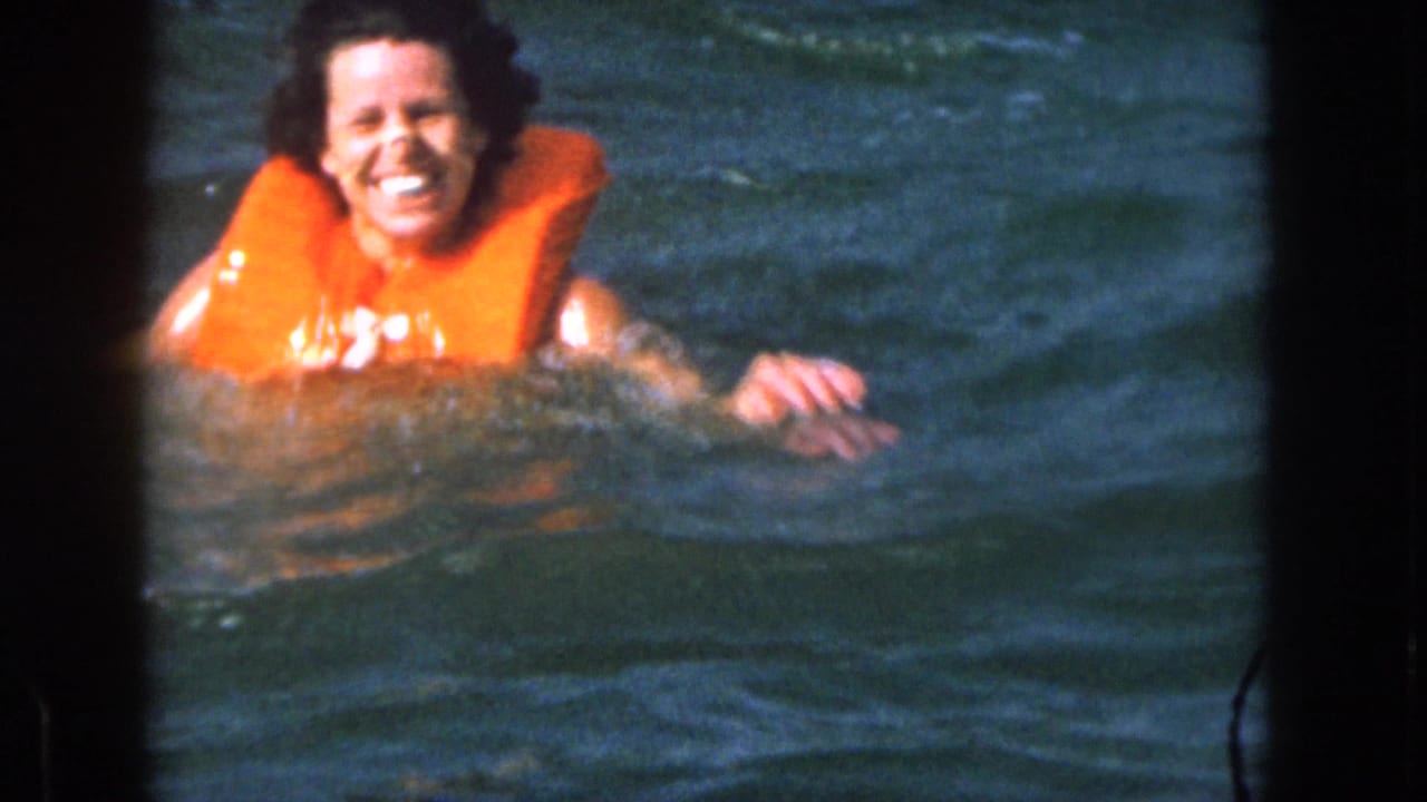 In 1961 Michigan home-movie footage, a smiling woman floats in lake water wearing a bright orange life vest.