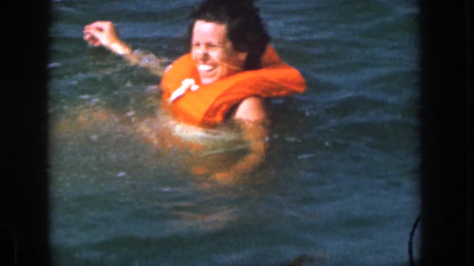 The woman laughs in the water with one arm raised, framed by the soft color and grain of 1961 Michigan 8mm footage.