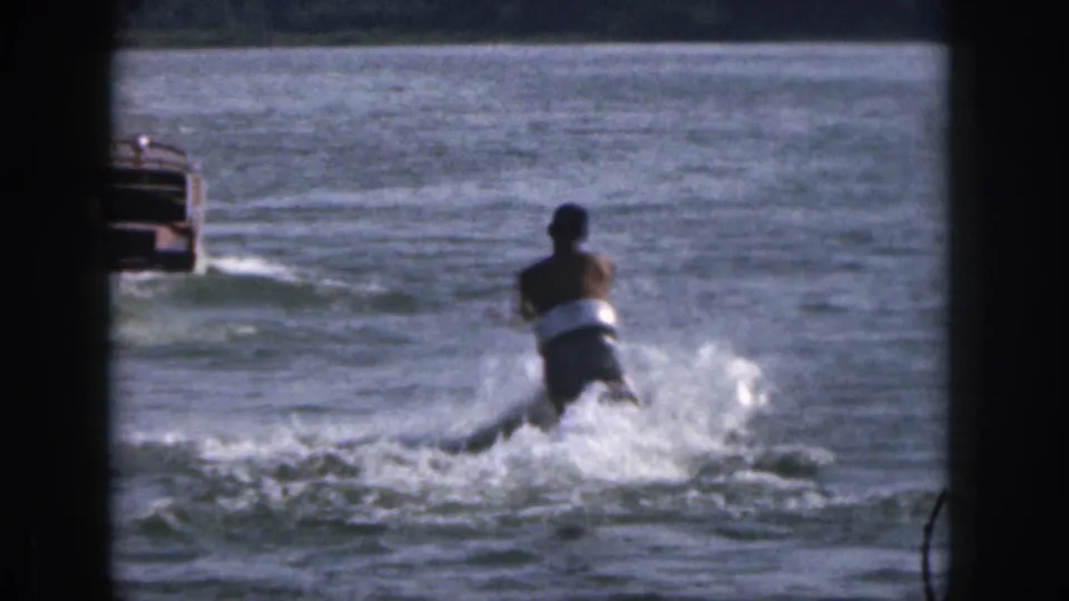 A distant skier follows a small boat across the lake in 1961 Michigan home-movie footage, with spray visible behind them.