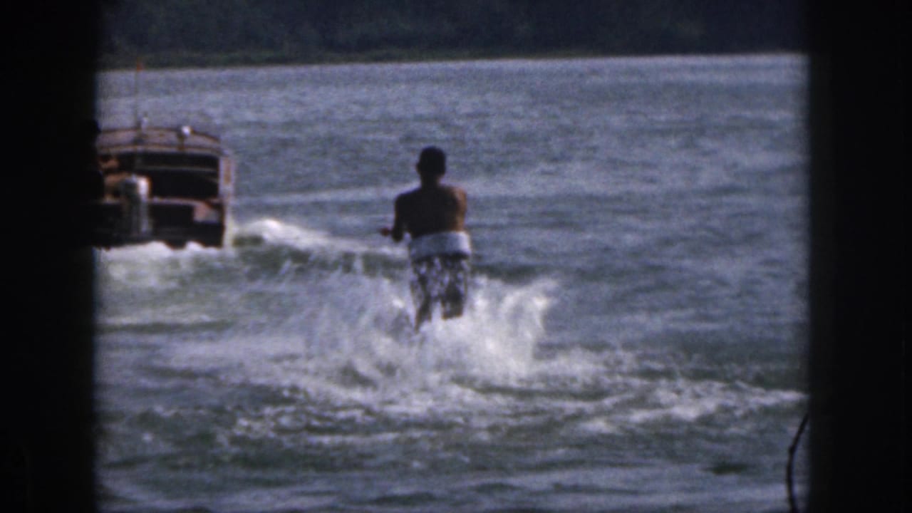 1961 Michigan home-movie frame showing a shirtless water skier behind a motorboat on a lake with white spray and trees in the distance.