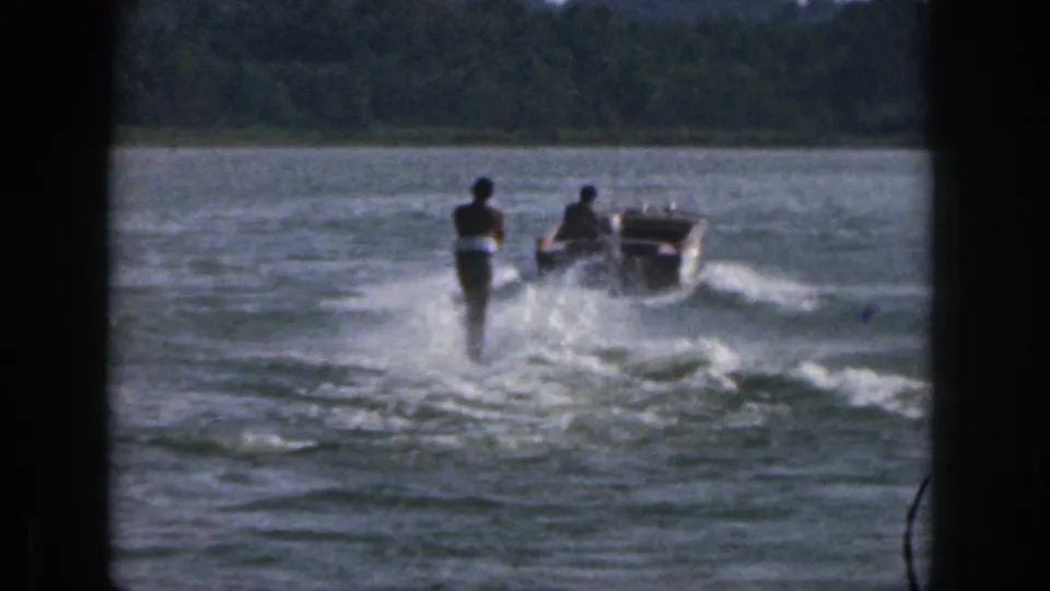 1961 Michigan 8mm frame showing a water skier behind a small boat, both moving away across the lake toward a wooded shore.
