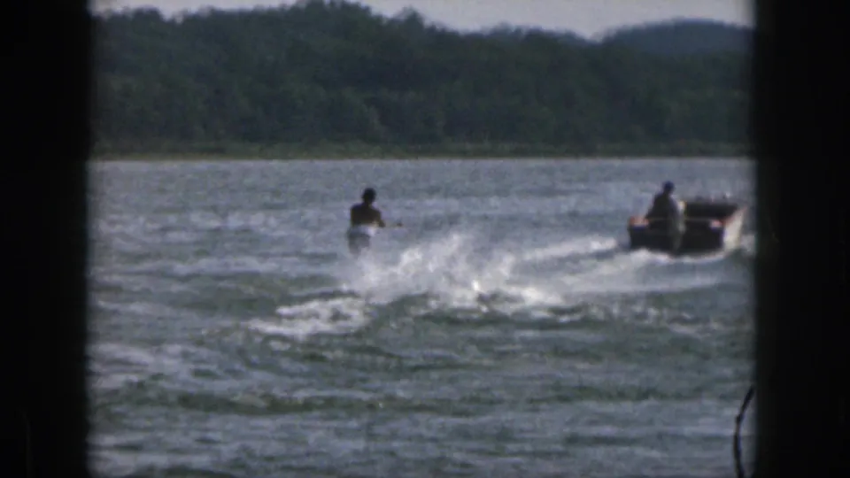 1961 Michigan home-movie frame with a distant skier and motorboat crossing a lake, framed by dark film borders and a wooded horizon.