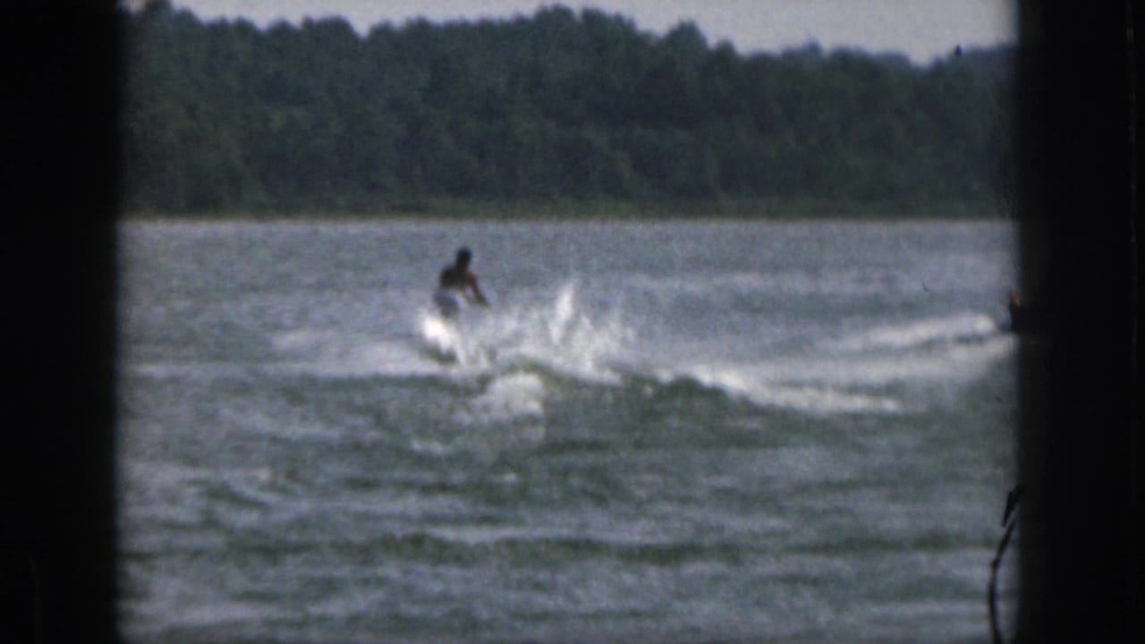 In 1961 Michigan home-movie footage, a distant water skier moves across a lake, throwing spray against a dark tree-lined shore.