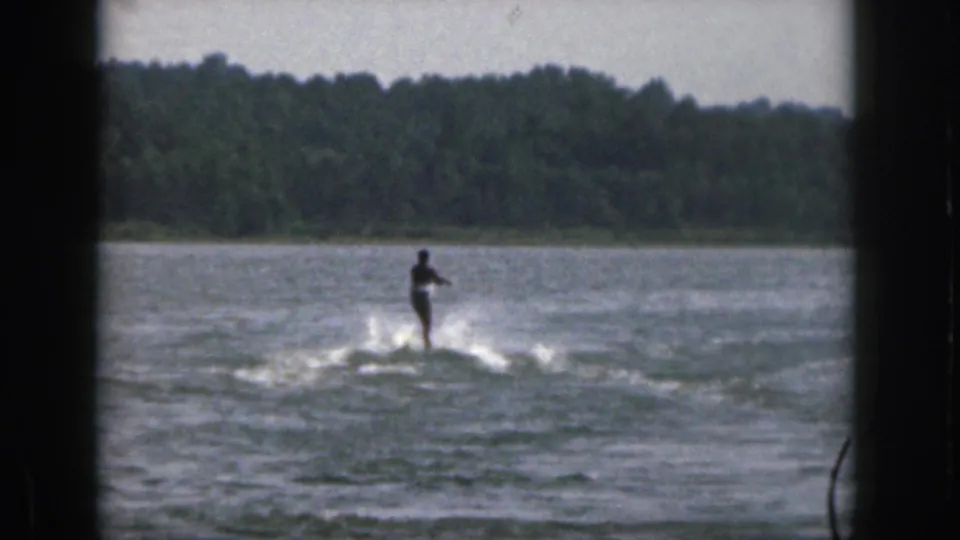 A distant water skier in the Michigan lake scene stands upright behind a tow line, with rippled water and trees beyond.