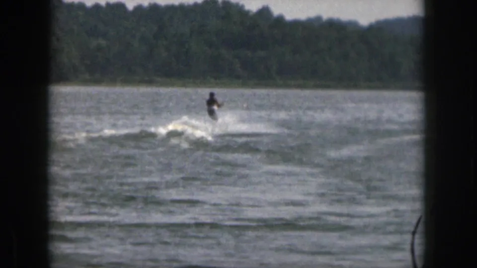 The 1961 Michigan home movie shows the skier farther from camera, framed by lake water, white wake, and a wooded shoreline.