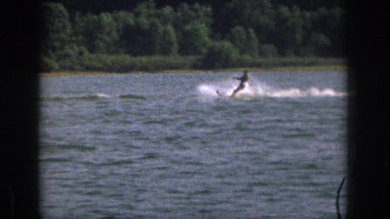 A distant skier in dark swimwear is pulled across a Michigan lake, with spray trailing over the rippled water.