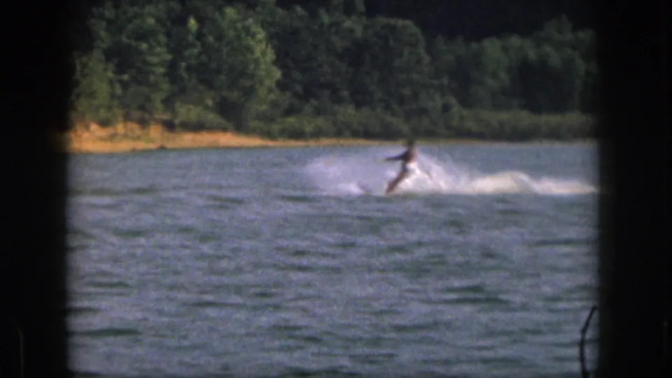 The 8mm frame shows the skier leaning against the pull of the rope, with white spray and a wooded bank behind.