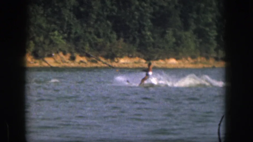 A distant water skier crosses the lake in front of a sandy bank and dense trees, leaving a bright trail of spray.
