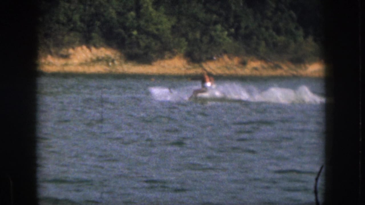 In 1961 Michigan 8mm footage, a distant shirtless man water skis across blue lake water with trees behind him.