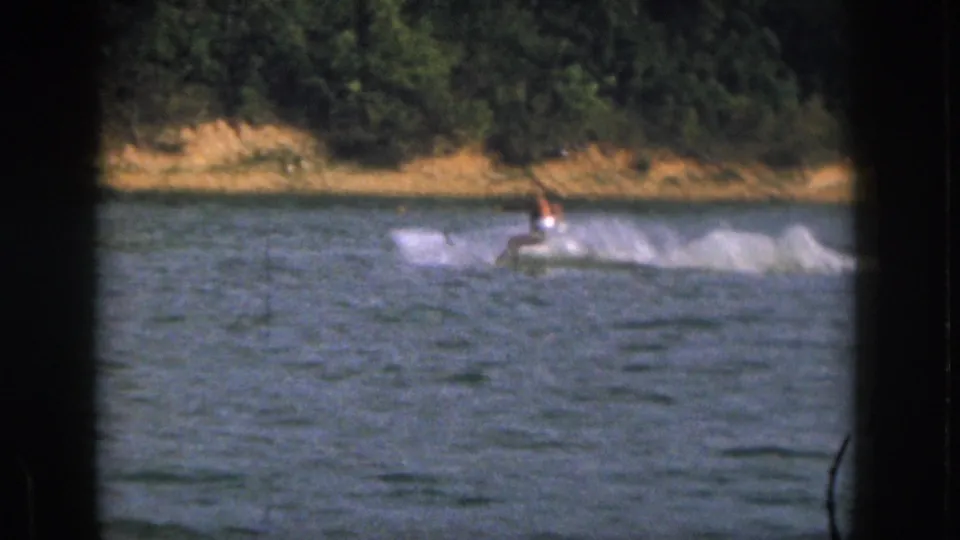 In 1961 Michigan 8mm footage, a distant shirtless man water skis across blue lake water with trees behind him.