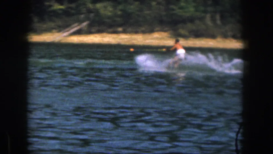 A shirtless man in white trunks water skis across the lake, with choppy blue water and a wooded bank behind him.