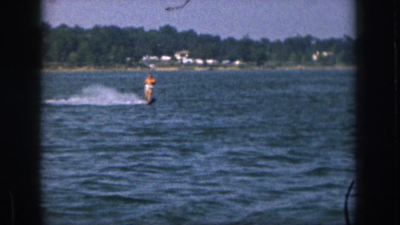 1961 Michigan home-movie frame showing a man in an orange shirt water skiing on a large lake, with trees and distant houses beyond the water.