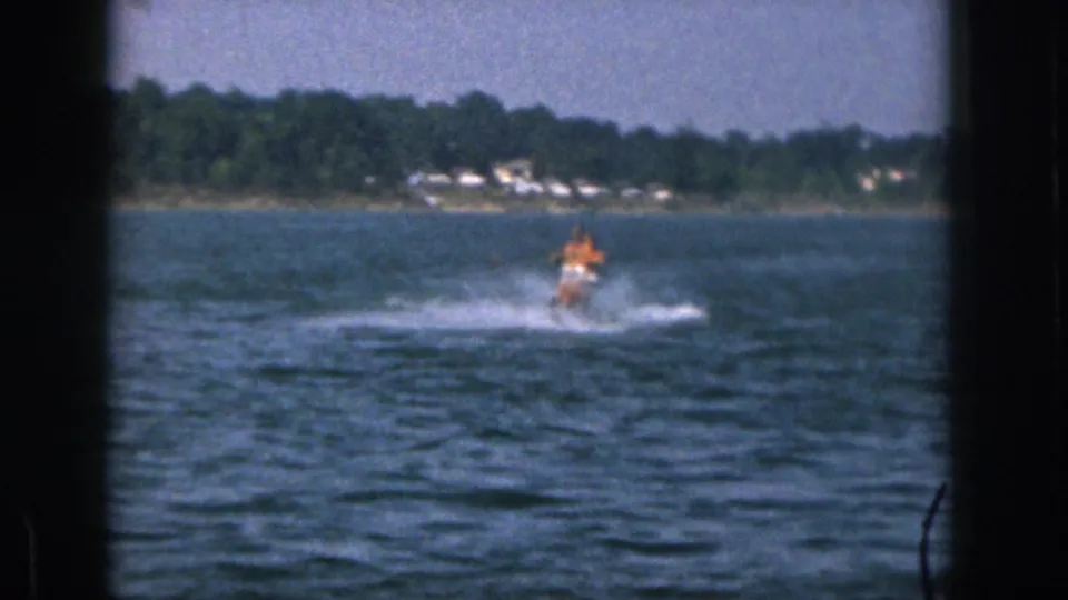 1961 Michigan home-movie view of a man on a single water ski moving toward the camera across a broad lake with a distant wooded bank.