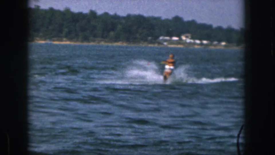 A 1961 Michigan lake recreation frame shows the distant water skier cutting across choppy water, with spray, trees, and small shoreline houses behind.