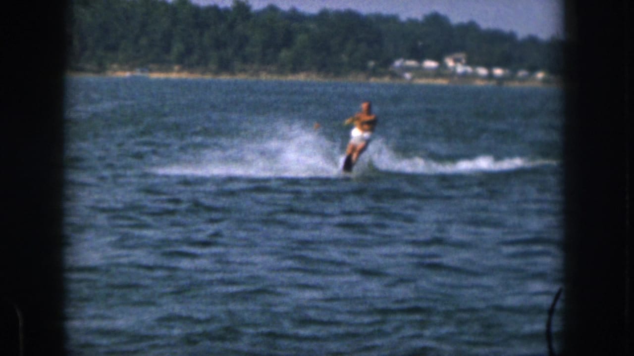 1961 Michigan home-movie frame of a shirtless man in white shorts water-skiing across a lake near a wooded shore.