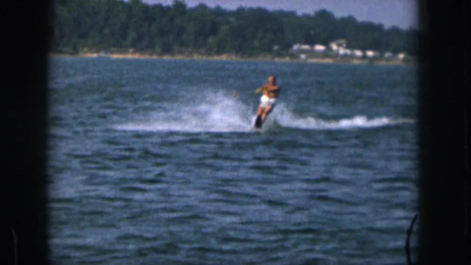 1961 Michigan home-movie frame of a shirtless man in white shorts water-skiing across a lake near a wooded shore.
