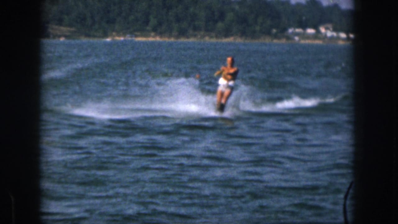 Water-skier gliding across blue lake water with spray and a tree-lined shore in back.