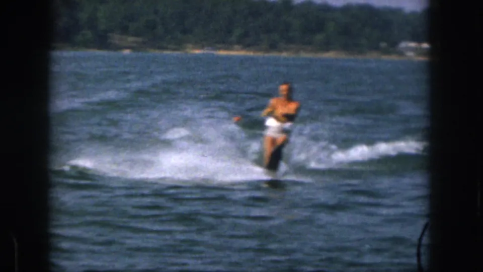 Closer 1961 Michigan lake frame with an adult man in white shorts water-skiing through choppy spray.