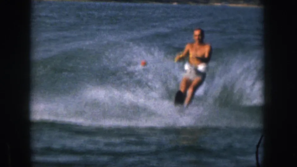 1961 Michigan home-movie frame of a shirtless adult man water-skiing close to camera with heavy lake spray.