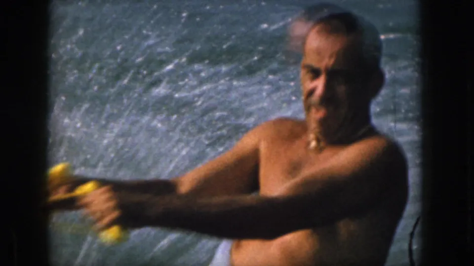 Close 8mm view of the water skier's face and arms as he holds a yellow tow handle against a spray-filled river background.