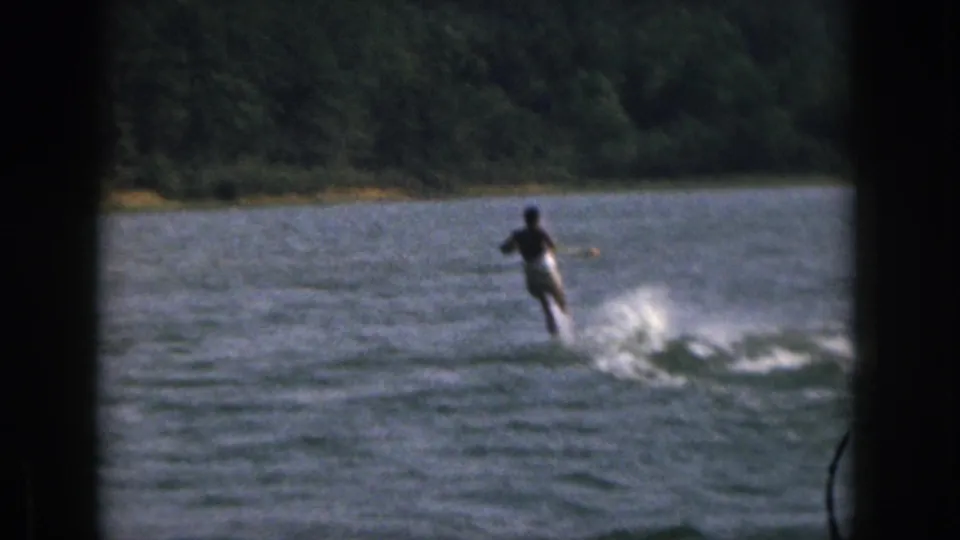 Wide 8mm frame from Michigan showing a distant adult water skier moving across the river with spray trailing and trees along the far bank.
