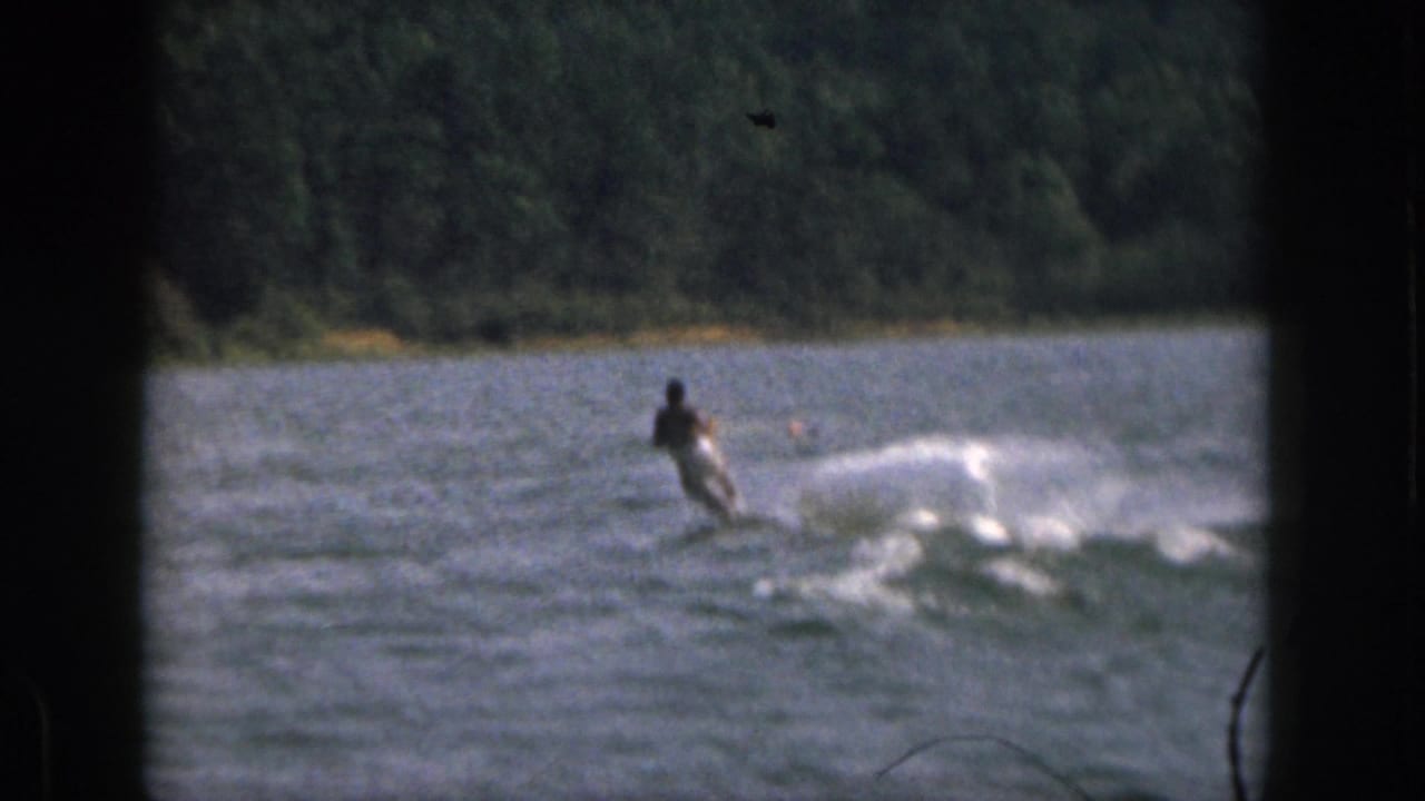 1961 Michigan 8mm frame showing a water skier moving across a lake, with wake spray and a forested shoreline beyond.