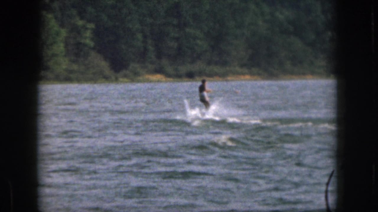 1961 Michigan home-movie frame with a distant water skier riding upright across open water near a tree-lined shore.