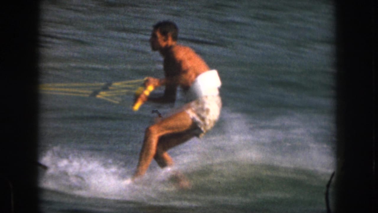 1961 Michigan home-movie frame showing a shirtless adult man balancing low on water skis while holding a yellow tow rope.
