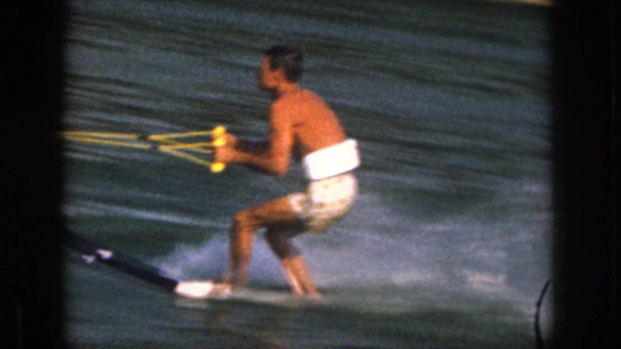 1961 Michigan frame of an adult man in patterned trunks and a white buoyancy aid skiing across lake water.
