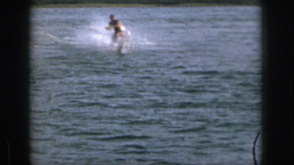 1961 Michigan home-movie frame showing the skier farther from camera on a lake, trailing a small spray wake.
