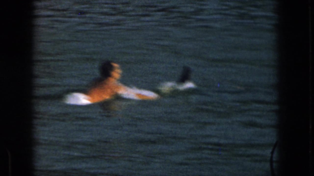 Shirtless adult man floating in a lake with water skis visible behind him.