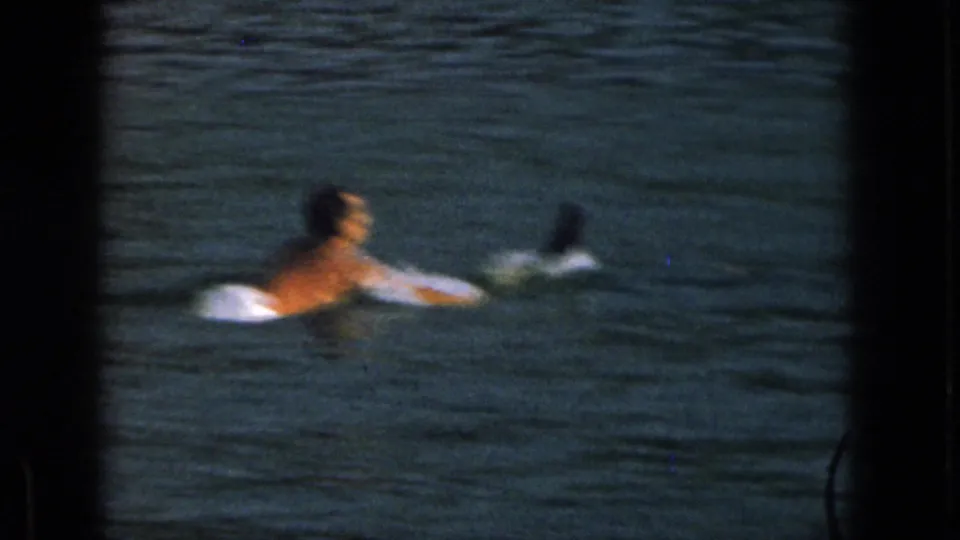 In this Michigan home-movie frame, a shirtless adult man floats in lake water with water skis trailing as he prepares to continue.