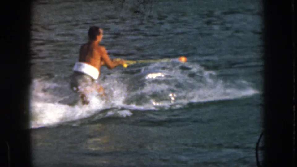 A shirtless adult man in swim trunks grips a tow rope as he starts across choppy Michigan lake water on skis.