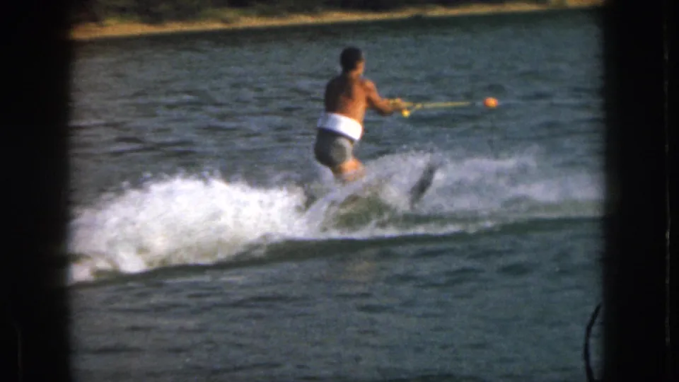 The adult skier stands more upright on the lake, holding the tow rope as a small wake and shoreline appear in the 8mm frame.