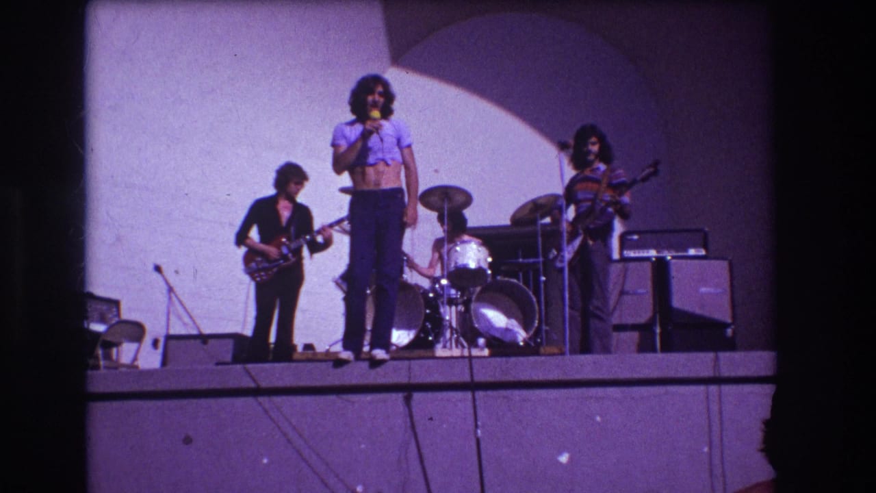 Four-piece band on stage with singer at microphone, guitarist, bassist, drummer, amplifiers, and white backdrop.