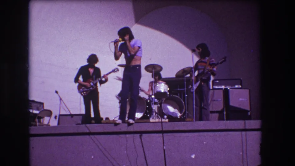 The 1973 Medford stage scene continues with the singer turned toward the microphone and the band visible against a plain backdrop.