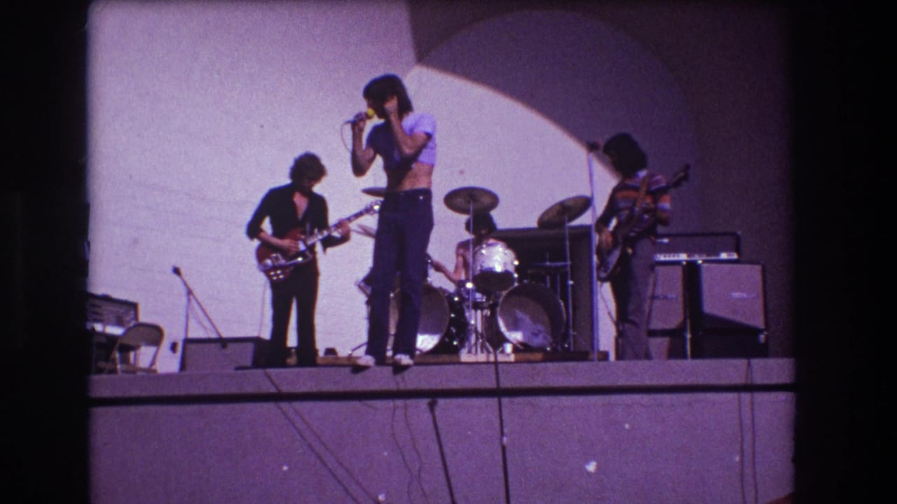 Rock band on a raised stage with singer, guitarist, drummer, bassist, amplifiers, and curved backdrop.